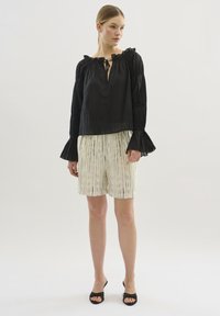 Schwarze, locker sitzende Bluse mit rüschigem Ausschnitt und langen, ausgestellten Ärmeln. Kombiniert mit cremfarbenen Shorts mit vertikalen schwarzen Streifen. Schwarze Sandalen.