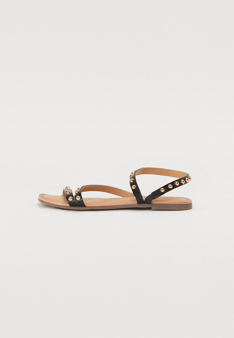 Sandales plates noires en cuir avec des accents cloutés dorés et un design de brides, dotées d'une semelle minimaliste et d'un bout ouvert.