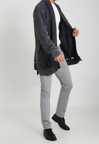 Lång, mörkgrå cardigan med en mjuk textur, öppen front, breda ärmar och sidoflikar, bärs över en vit skjorta och grå byxor.