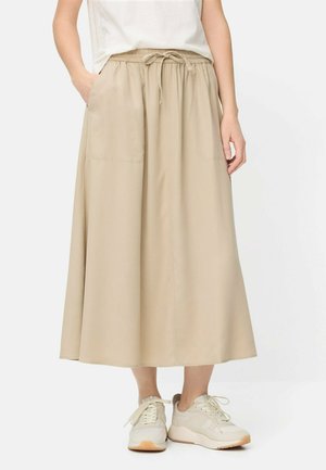 Beżowa midi spódnica z elastycznym pasem i sznurkiem, noszona z białymi trampkami i białym włożonym do środka topem, ręce modelki częściowo w kieszeniach.