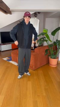 Juoda liemenė be rankovių ant tamsaus džemperio, derinama su laisvomis juodomis kelnių ir sidabriniais batais. Vidaus aplinka su medinėmis grindimis ir vazonine augalu.