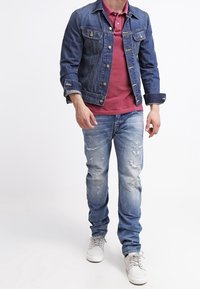 Hombre caminando con una chaqueta de mezclilla azul, camisa polo roja, jeans azules desgastados y zapatillas blancas frente a un fondo blanco liso.