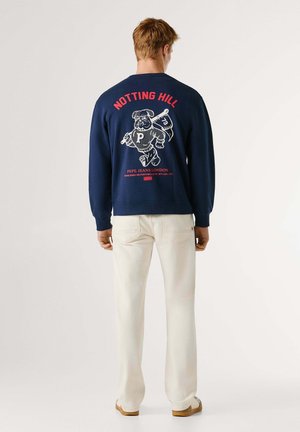 Marineblauer Sweatshirt mit Bärengrafik und rotem Text auf der Rückseite. Verfügt über gerippte Bündchen und Saum. Kombiniert mit hellen Hosen und Schuhen.