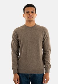 Bruine crewnecktrui van een zachte stof, met lange mouwen en een geribbelde zoom. Gepaard met blauwe denim jeans staat het model voor een witte achtergrond.