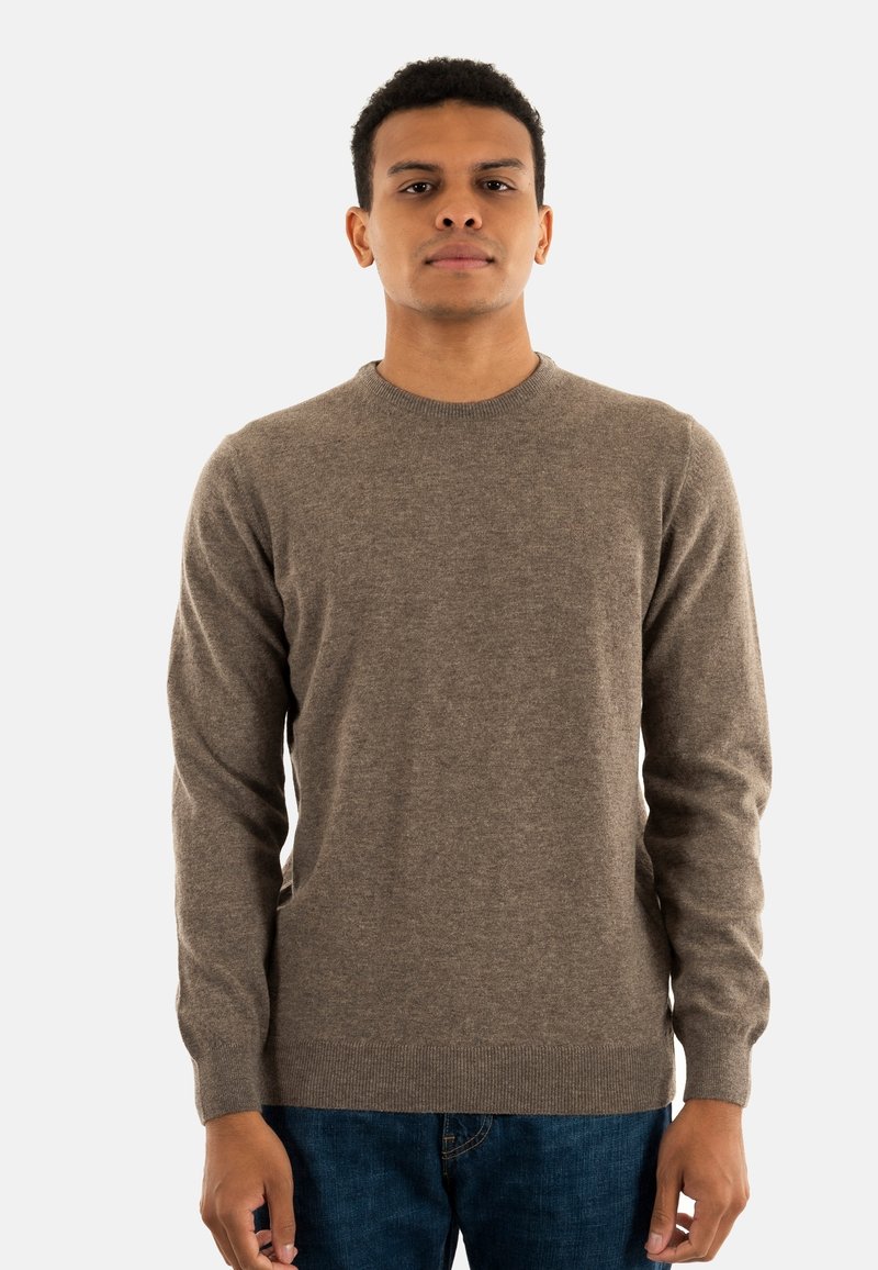 Bruine crewnecktrui van een zachte stof, met lange mouwen en een geribbelde zoom. Gepaard met blauwe denim jeans staat het model voor een witte achtergrond.