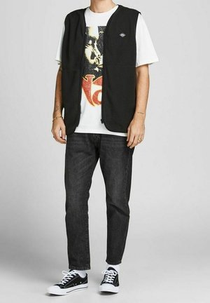 Hombre que lleva camiseta blanca con gráfico, chaleco polar negro con cremallera, vaqueros negros, calcetines blancos y zapatillas negras, de pie frente a un fondo gris.