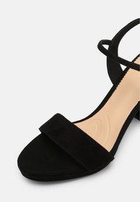 Sandale à talon en suede noir avec une large bride sur l'avant, une fine bride autour de la cheville et une semelle intérieure lisse beige. Le talon est de hauteur modérée.