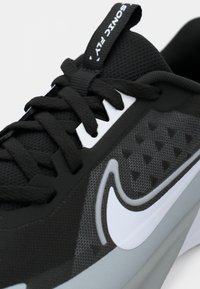 Fekete sportcipő hálós felsőrésszel, lapos fűzőkkel, gumis talppal és egy feltűnő fehér logóval. Figyelemre méltó textúrált minták az oldalon.