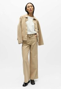 Bēša oversized jaka un atbilstošas platām kājām bikses ar aukliņu jostasvietā, kombinētas ar baltu kreklu un melniem apaviem. Gluda auduma tekstūra.