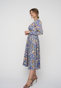 Geprinte jurk in blauw met bloemen- en paisleypatronen. Lange mouwen, getailleerde corsage, uitlopende rok. Draagt met gouden sandaaltjes met hakken.