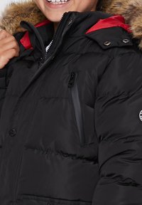 Veste rembourrée noire avec une doublure intérieure rouge, capuche avec bordure en fourrure, fermeture éclair à l'avant et plusieurs poches zippées. Matériau doux et texturé.