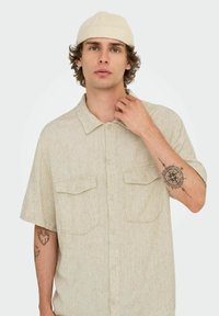 Kortærmet beige skjorte med to brystlommer, har et struktureret stof, frontknapper og en afslappet pasform. Sammen med en beige kasket.