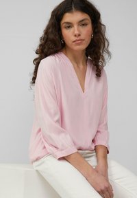 Blouse rose clair avec un col en V, des manches trois-quarts et des poignets froncés. Tissu lisse et coupe ample. Portée avec un pantalon blanc.