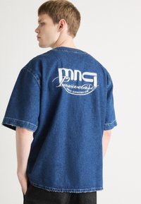 Denim oversized skjorte i mørkeblå med korte ermer. Har en hvit brodert logo på baksiden med "MNC Paradise Disco Dancing Club."