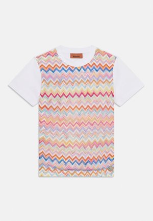 Biała koszulka z krótkim rękawem z kolorowym wzorem dzianym w zygzak na przodzie oraz małym, okrągłym srebrnym logo blisko dolnego brzegu.