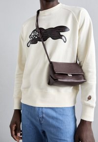 Felpa beige con design grafico scuro sul davanti, abbinata a una borsa a tracolla in pelle marrone strutturata. I jeans in denim blu completano il look.