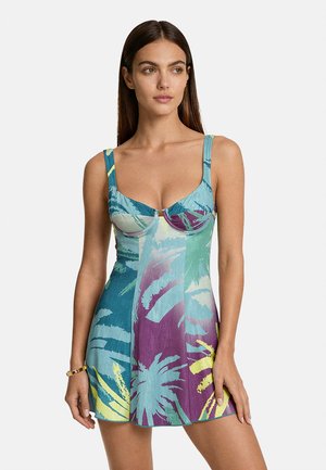 Donna con capelli lunghi castani che indossa un abito senza maniche con stampa tropicale multicolore, corpetto aderente e gonna svasata, in piedi su sfondo bianco.