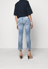 Persona con jeans cortos de color azul claro con ligero desgaste, una blusa negra holgada y sandalias de tacón alto blancas con tiras, de pie de espaldas.