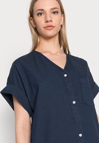 Camisa azul marinho de manga curta com decote em V, botões frontais e um bolso no peito. Fabricada em tecido leve com mangas arregaçadas para um ajuste descontraído.