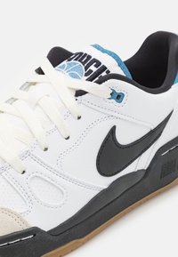 Zapatilla blanca con swoosh negro, de cuero texturizado y suela de goma. Presenta cordones crema y un acento azul en la lengua con la marca.