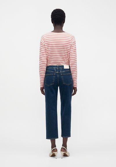 Femme debout de dos, portant un chemisier à manches longues rayé rose et blanc, un jean court bleu foncé et des sandales beiges à talons hauts.
