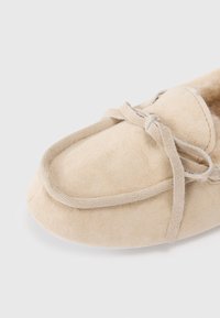 Pantoufle en suède beige avec un bout rond, un bord cousu et un détail en nœud en tissu sur le dessus. Texture lisse et doublure douce visibles.