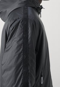 Chaqueta de rompevientos negra con capucha y una cinta de marca vertical que recorre el brazo, con detalles de cordón en la capucha y el bolsillo.