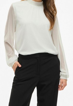 Femme portant un chemisier blanc à manches longues avec des poignets à volants et un pantalon noir taille haute, la main dans la poche, debout devant un fond uni.