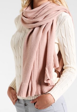 Femme portant un pull en maille torsadée crème et une grande écharpe douce rose pâle à bords frangés, les mains dans les poches d'un pantalon gris clair.