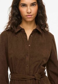 Pruun sametkangast triiksärkkleit, millel on krae, nööbitav esikülg ja vööümbermõõdu seotav pael. Omaduseks vertikaalne sooniline tekstuur ja sobivad pruunid nupud.