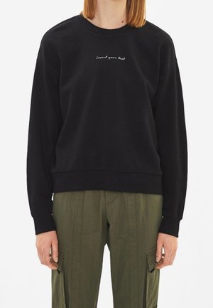 Sweatshirt noir avec un col rond et des poignet côtelés, arborant un petit détail textuel blanc « insérez votre texte » sur la poitrine. Associé à un pantalon cargo vert olive.