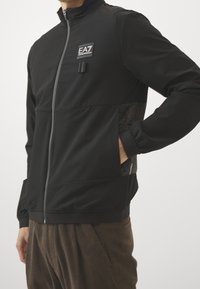 Svart sportjacka med hög krage, dragkedja framtill, texturerade sidopaneler och elastiska ärmslut. Har en logotypapplikation på bröstet.