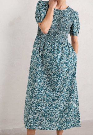 Nainen, joka pukeutunut tummanvihreään kukalliseen midimekkoon, jossa on rypytetty yläosa, liehulakit ja sivutaskut, yksinkertaista vaaleaa taustaa vasten.