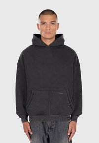 Mand med kort hår, iført en mørkegrå hættetrøje og slidte jeans, der står mod en ensfarvet lys baggrund.