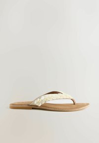 Sandale plate couleur beige avec des embellissements blancs en forme de pétales sur la lanière en tissu au niveau des orteils, avec un design tong et une bordure de semelle cousue.