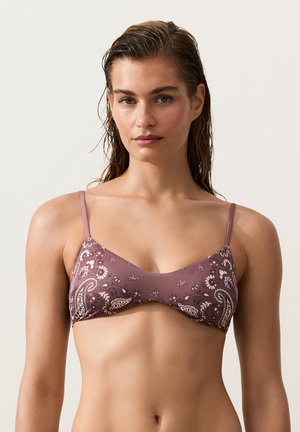 Młoda kobieta z mokrymi włosami, ubrana w górę bikini w kolorze mauve z wzorem paisley na jednolitym jasnym tle.