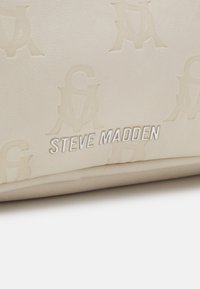 Beżowa torebka z faux skóry z wytłoczonym wzorem logo oraz napisem "STEVE MADDEN" w kolorze białym. Gładka tekstura, prostokątny kształt.
