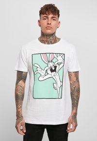 T-shirt bianco in cotone con un personaggio dei cartoni animati con grandi orecchie, delineato in nero su uno sfondo teal, inserito all'interno di una cornice rettangolare.