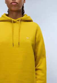 Žlutá mikina s kapucí z bavlněné směsi, obsahující stahovací šňůrky, malé vyšívané logo na hrudi a měkký, texturovaný povrch.