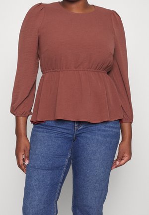 Femme portant un top à basque à manches longues couleur rouille avec une taille élastique et un jean en denim bleu sur un fond uni.