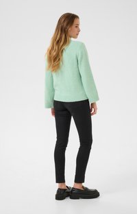 Světle zelený svetr s texturovaným pletením a žebrovanými manžetami, kombinovaný s černými upnutými džíny a černými platformovými loafers. Model otočený zády.