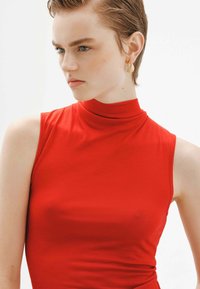 Top rojo sin mangas con un escote alto hecho de una tela suave. El modelo lleva un pendiente dorado en forma de gota y el cabello corto peinado de manera ordenada.