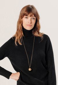 Zwarte coltrui met geribbelde manchetten, vergezeld van een lange gouden hanger met een bolvormige bedel. Eenvoudig en elegant ontwerp.