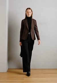 Hnědý blazer přes černý rolák, v kombinaci s černými širokými kalhotami. Černé boty na podpatku a černá kabelka dotvářejí celkový vzhled.