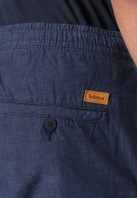 Navy blue shorts made from a textured fabric, featuring an elastic waistband, button detail, and a tan Timberland logo patch.