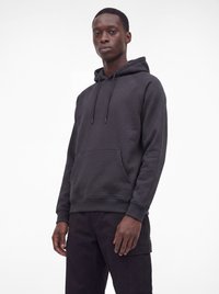 Un homme portant un sweat à capuche noir avec une poche avant et des cordons, associé à un pantalon cargo noir, debout devant un fond blanc uni.