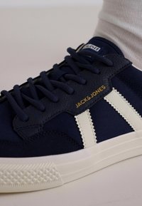 Námořnické plátěné tenisky s krémovými akcenty, které mají texturovanou gumovou podrážku, šněrovací design a zlaté logo "JACK & JONES" na boku.
