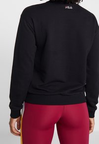 Felpa nera in pile con orlo e polsini a costine. Piccolo logo ricamato sul colletto posteriore. Abbinato a leggings rossi.