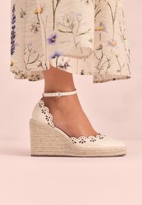 White wedge heel shoe with a woven jute platform, scalloped cutouts, and a thin ankle strap. Smooth leather surface, featuring floral accents.
