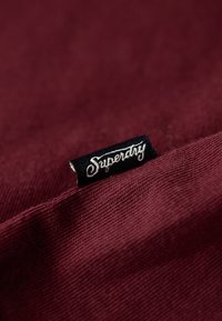 Zwarte stoffen label met witte "Superdry" tekst op een bordeaux geknoopte kledingstuk, waarbij de textuur en stiksels van de stof zichtbaar zijn.